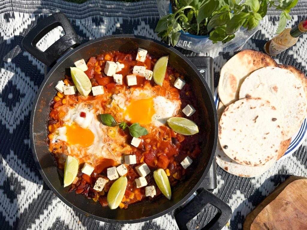 A skillet with Mexican shakshuka on a camping stove, filled with spicy tomato sauce, black beans, corn, eggs, feta, and lime, served with warm tortillas during outdoor cooking.